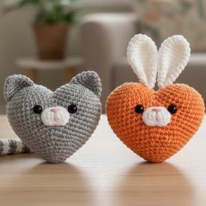May include: Two handmade crochet heart-shaped toys. One is gray and cat-shaped, with a striped tail. The other is orange with white bunny ears. Both have black eyes and pink noses, perfect for nursery decor.