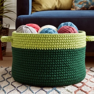 May include: A handmade, two-tone crochet basket in dark green and light green, filled with colorful yarn balls. The basket has two small handles and sits on a patterned rug, suggesting a craft or home decor theme.