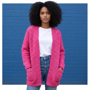 May include: A bright pink, open-front crochet cardigan with two front pockets. The cardigan is worn over a white t-shirt and blue jeans. The background is a solid blue brick wall. The cardigan is a medium weight and appears to be handmade.