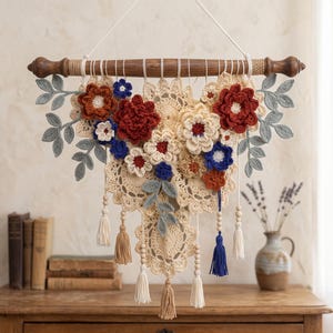 May include: A wall hanging featuring a dark wooden rod with white cord, adorned with crocheted cream-colored doilies, layered with red, rust, and blue crocheted flowers and grey leaves. The piece includes hanging wooden beads and cream and blue tassels.