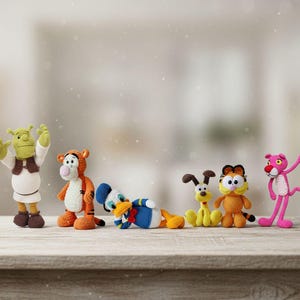 Op de afbeelding: Een verzameling van zes gehaakte figuren, waaronder een groene oger, een oranje tijger, een blauw-witte eend, een gele hond, een oranje cyperse kat en een roze panter, allemaal op een rij geplaatst op een houten ondergrond.