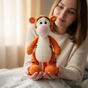 May include: A crocheted tiger toy in orange and white, with black stripes and a pink nose. The toy is held up, showing its features, including orange ears. The background is soft and out of focus.