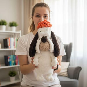 Puede incluir: Un gran peluche de perro hecho a mano con ganchillo. El perro es principalmente blanco con orejas marrones, un hocico marrón y un postizo naranja texturizado. El peluche se sostiene frente a una persona, mostrando su tamaño y detalle.