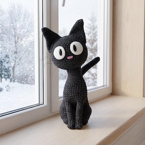 Puede incluir: Figura de gato negro de ganchillo hecha a mano con grandes ojos blancos y una nariz rosa, sentado en un alféizar. El gato tiene un brazo levantado. El fondo muestra una escena invernal nevada.