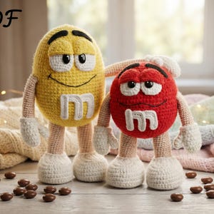 May include: Two crocheted figures, resembling M&M's sweets. One is yellow with a white "M", the other is red with a white "M". Both have arms, legs, and smiling faces. The figures are on a wooden surface with scattered chocolate sweets.