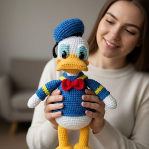 May include: A handmade crochet doll of Donald Duck, featuring a blue sailor shirt, a red bow tie, and a blue hat. The doll has a yellow beak and feet, and white accents on the head and hands. The doll is approximately 12 inches tall.