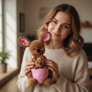 May include: A brown crochet kangaroo with pink inner ears and a pink pouch, holding a smaller kangaroo. The handmade stuffed animal has a sweet expression and is a charming toy.
