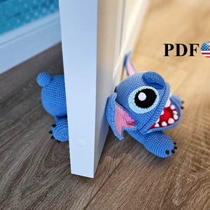 May include: A blue crocheted Stitch doorstop, with large eyes, pink ears, and a wide open mouth revealing red and white teeth. The doorstop is positioned next to a white door frame on a wooden floor. The text "PDF" is visible.