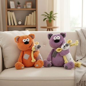 Könnte beinhalten: Zwei gehäkelte Teddybären, einer orange und einer lila, sitzen auf einem cremefarbenen Sofa. Jeder Bär hält ein kleines graues Mausspielzeug mit gelben Akzenten. Die Bären haben große Augen und lächeln. Im Hintergrund sind ein Bücherregal und ein Fenster zu sehen.