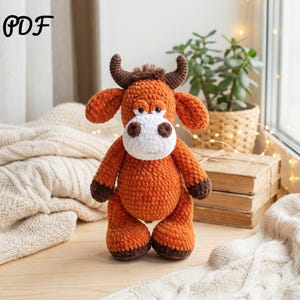Peut inclure: Un taureau au crochet orange fait à la main avec des cornes, des sabots et un visage blancs. L'animal en peluche a une expression amicale et se tient sur une surface en bois. Le mot "PDF" est dans le coin supérieur gauche.