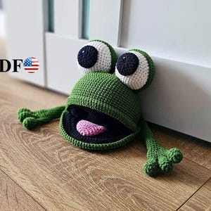 Puede incluir: Una rana verde de ganchillo hecha a mano con grandes ojos blancos y negros, una boca abierta que revela una lengua rosa y brazos extendidos. La rana está colocada sobre un suelo de madera, apoyada contra una puerta blanca. La palabra "PDF" es visible.