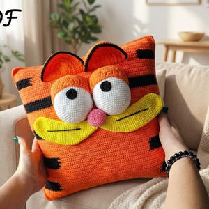 Könnte beinhalten: Ein orangefarbenes Häkelkissen mit einem Cartoon-Katzen-Gesicht, schwarzen Streifen, großen weißen Augen und einem gelben Schnurrbart. Es hat eine rosa Nase und den Text "PDF" in Schwarz in der oberen linken Ecke.