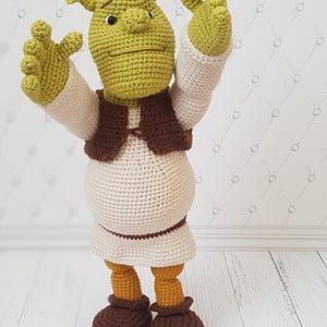 Puede incluir: Una muñeca de ganchillo hecha a mano de un personaje ogro verde con una cabeza grande, orejas en forma de trompeta y una nariz ancha. La figura viste una túnica color crema, un chaleco marrón, un cinturón marrón y botas marrones.