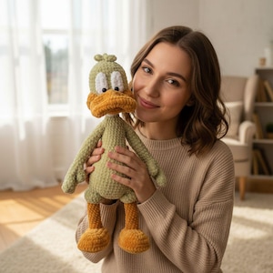 Puede incluir: Un muñeco de pato de ganchillo hecho a mano, verde y naranja. El pato tiene un pico y patas grandes de color naranja, con ojos blancos y una cabeza verde. El muñeco lo sostiene una persona que lleva un jersey beige.