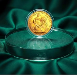 May include: A gold coin featuring a rooster design, encased in a clear protective holder. The coin is displayed on a clear, circular stand against a backdrop of dark green fabric. The coin has the text "LIBERTE EGALITE FRATERNITE".