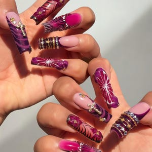 Könnte beinhalten: Ein Set langer, verzierter Kunstnägel. Designs umfassen Kreuze, Blumenmuster und geometrische Formen in Pink, Lila und Gold. Einige Nägel weisen Strasssteine und andere dekorative Elemente auf.