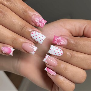 Puede incluir: Uñas manicuradas con diversos diseños. Algunas uñas tienen una manicura francesa con base rosa, otras son blancas con lunares rosas. Dos uñas presentan un diseño de flor rosa en 3D. Las uñas están bien cuidadas y pulidas.