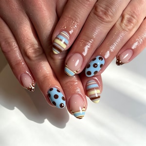 May include: Manicured fingernails featuring an almond shape with a mix of designs. Some nails display horizontal stripes in light blue, yellow, and brown, while others feature light blue polka dots on a brown base or brown tips with white dots.