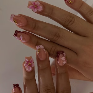 May include: Artificial nails in a square shape, painted nude with burgundy and pink French tips. Some nails have 3D pink flower designs and small gold embellishments. A manicure with a floral theme.
