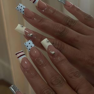 以下が含まれることがあります： フレンチマニキュアのデザインが施された、長くて四角いアクリルネイル。一部の爪は、シルバーの装飾が施された白いチップが特徴で、他の爪は、ライトブルーのドット柄または、バーガンディ、白、ライトブルーのストライプ柄です。爪は光沢のある仕上がりです。