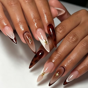 May include: Long almond-shaped nails featuring a mix of glossy dark brown tortoiseshell patterns, solid brown tips, and nude bases. Some nails are accented with white French tips, small gold metallic spheres, and delicate gold starburst charms.