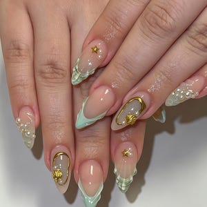 Puede incluir: Uñas en forma de almendra con un diseño de punta francesa en verde menta claro y acabado ondulado. La manicura está decorada con pequeñas perlas blancas, diminutas estrellas doradas y marcos ovalados dorados con conchas marinas en miniatura.