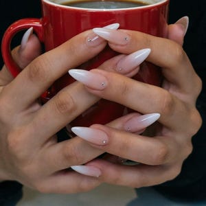Pode incluir: Uma caneca de cerâmica vermelha cheia de um líquido escuro, segurada por mãos com unhas longas e pontiagudas em estilo ombre. As unhas fazem a transição de uma base nude para uma ponta branca, com pequenos enfeites prateados.