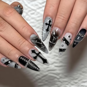 May include: A set of long, pointed fingernails with a gothic theme. The nails feature a variety of designs, including black crosses, chains, and star accents, set against a palette of black, white, and gray.