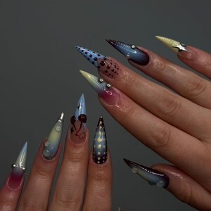 May include: Close-up of long, pointed acrylic nails with diverse designs. The nails display polka dots, cherry motifs, and gradient colour schemes. Some nails are decorated with small, silver embellishments.