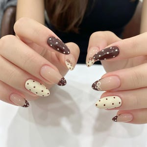 May include: Almond-shaped nails featuring a mix of dark brown and cream polish. Some nails are solid with white or brown polka dots, while others have a nude base with brown or cream tips, accented with small hand-painted bows at the cuticle line.