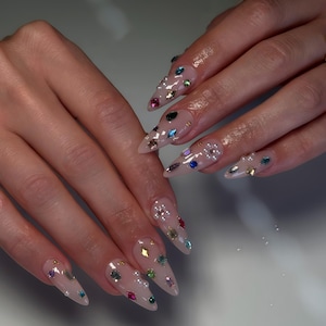Puede incluir: Primer plano de uñas largas y puntiagudas con una base transparente y adornos de gemas coloridas y brillantes. Las gemas son de varias formas y tamaños, incluyendo redondas, cuadradas y en forma de lágrima, en tonos rojos, azules, verdes y dorados.