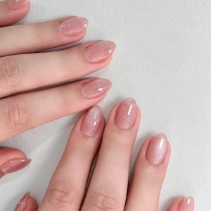 May include: Oval-shaped fingernails featuring a sheer, light pink polish with a subtle glitter finish. The nails are groomed to a smooth, rounded edge, showcasing a glossy, semi-transparent appearance that catches the light across the surface.