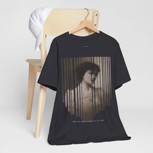 May include: Dark grey t-shirt with a sepia-toned portrait of a woman and vertical bar overlays. The shirt has the text "HOLD THY WORD AS DEARLY AS THY NAME". A white baseball cap rests on a wooden chair.