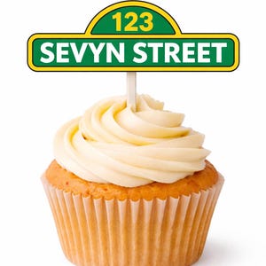 May include: A cupcake with vanilla frosting and a decorative sign that reads "123 Sevyn Street". The sign is green with yellow accents and is attached to a white stick. The cupcake is in a paper liner.