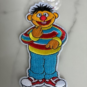May include: Embroidered patch featuring a cartoon character with yellow skin, a red nose, and black hair. The character wears a striped shirt in red, blue, and yellow, blue pants, and red and white sneakers. The patch has a white border.