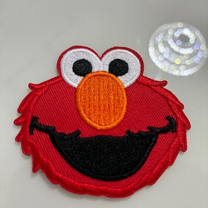 May include: A red embroidered patch featuring the face of a cartoon character with white eyes, black pupils, an orange nose, and a black smiling mouth. The patch has a textured appearance and is likely for clothing or accessories.