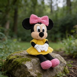 Może przedstawiać: Szydełkowa lalka myszki z czarnymi uszami, różową kokardą i żółtą sukienką. Twarz lalki jest beżowa z czarnymi oczami i czarnym nosem. Lalka siedzi na skale pokrytej mchem. Stopy i dłonie są różowe i białe.