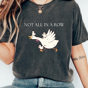 May include: Dark gray t-shirt with the phrase "NOT ALL IN A ROW" printed in white. The shirt features a cartoon illustration of a white goose with three yellow ducklings riding on its back. The goose is running with its wings spread.