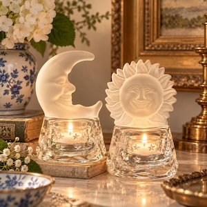 May include: Two glass tea light candle holders featuring frosted glass toppers. One topper is shaped like a crescent moon with a face, and the other is a sun with a face. The clear glass bases are decorated with small star patterns and hold lit candles.