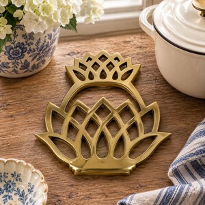 May include: A gold-toned metal trivet featuring a symmetrical, openwork design that resembles a stylized pineapple or floral motif with pointed, petal-like segments arranged in a tiered, lattice pattern with a smooth, reflective finish.