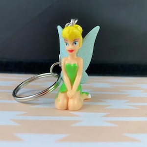 Könnte beinhalten: Ein Tinkerbell-Schlüsselanhänger. Die Figur kniet, trägt ein grünes Kleid und hellblaue Flügel. Der Schlüsselanhänger hat einen silbernen Ring.