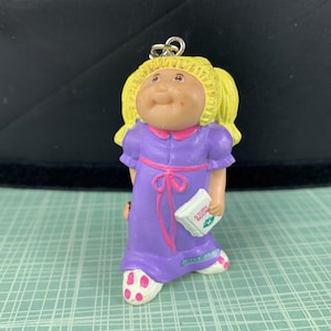 May include: A Cabbage Patch Kids doll figurine with blonde hair, wearing a purple dress with a pink bow. The doll is holding a small book that says "Story Book". The doll's feet are wearing white socks with pink polka dots.