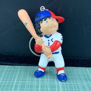 Puede incluir: Un llavero con temática de béisbol que presenta una figura de muñeca con uniforme de béisbol. La figura lleva una gorra azul y roja, un uniforme blanco con detalles rojos y zapatos azules. Sostiene un bate de béisbol de madera. El llavero está unido a un anillo plateado.