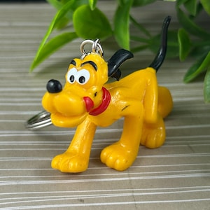 May include: A yellow Pluto keyring with a red collar, black ears, and a black tail. The cartoon dog is in a walking pose, with a silver keyring attached. Green foliage is visible in the background.