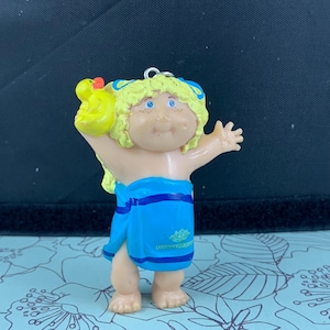May include: A Cabbage Patch Kids figurine of a child wrapped in a blue towel with blue stripes. The child has blonde hair, blue eyes, and is holding a yellow rubber duck. The figurine is a collectible item.