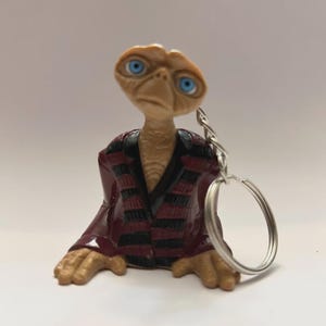 May include: A keychain featuring a figurine of the E.T. character. The figurine has a tan head and arms, blue eyes, and is wearing a burgundy jacket with black stripes. The keychain ring is silver.