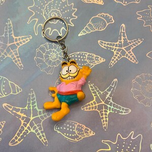 May include: A Garfield keychain featuring the cartoon cat in a pink shirt, blue shorts, and yellow legs. The keychain is attached to a silver ring. The background is a light blue pattern with starfish and seashell designs.