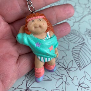 May include: A vintage Cabbage Patch Kids keyring featuring a doll in athletic attire. The doll has red hair, a pink headband, and is wearing a teal top with star accents, striped shorts, pink leg warmers, and purple shoes. The keyring has a silver chain.