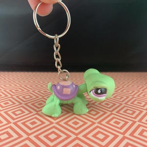 May include: A green turtle keychain with a purple shell and a silver keyring. The turtle has a detailed face with pink and black accents. The keychain is held up by a hand against a patterned background.