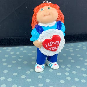 Op de afbeelding: Een Cabbage Patch Kids-beeldje met rood haar, een blauwe overall en een witte kraag. Het beeldje houdt een hartvormig bord vast met de tekst "I Love You" in rode letters. Het beeldje draagt witte schoenen met blauwe accenten.